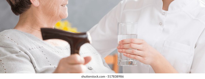 Caregiver proffering a glass of water for elderly woman with a hand on a cane - Shutterstock ID 517838986