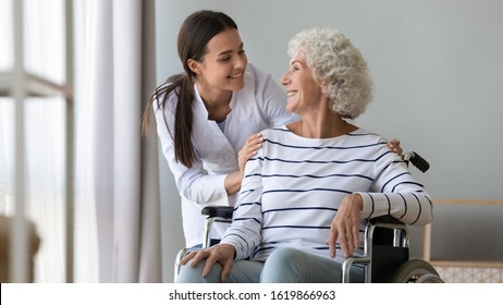 Caregiver Supporting Happy Disabled Older Woman Sitting In Wheelchair Close Up, Touching Shoulders, Expressing Care And Love, Smiling Nurse Wearing White Uniform And Mature Patient Having Fun