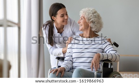Caregiver supporting happy disabled older woman sitting in wheelchair close up, touching shoulders, expressing care and love, smiling nurse wearing white uniform and mature patient having fun