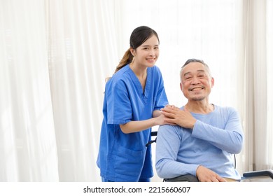The Caregiver Therapist Stands With An Asian Senior Sitting In A Wheelchair And Touches Their Hands On Senior Shoulder Together. The Nursing Home Facilitates A Support Group. Image With Copy Space