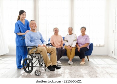 The Caregiver Therapist Stands With An Asian Senior Sitting In A Wheelchair With A Group Of Senior Women Sitting On A Sofa In The Background. The Nursing Home Facilitates A Support Group.
