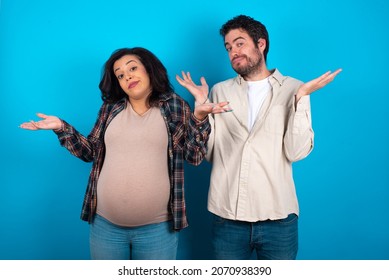 Careless Attractive Young Couple Expecting A Baby Standing Against Blue Background Shrugging Shoulders, Oops.