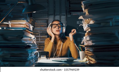 Careless lazy female employee sitting at office desk and listening to music instead of working, she is surrounded by piles of paperwork