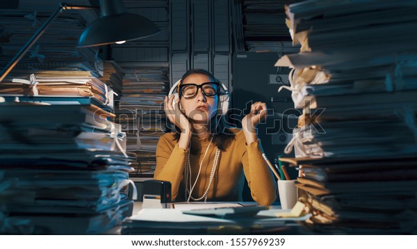 Careless lazy female employee sitting at office\
desk and listening to music instead of working, she is surrounded\
by piles of paperwork