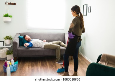 Careless And Slob Man Lying On A Couch And Watching TV While His Wife Does All The Housework At Home