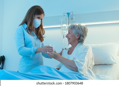 Carer Or Young Daughter Wearing A Face Mask Giving A Smiling Elderly Woman Patient Medication And A Glass Of Water As She Sits In A Hospital Bed