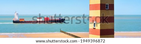 Cargo container ship sailing in the sea. Coast of Vlissingen, the Netherlands. Dunes and a striped lighthouse. Freight transportation, nautical vessel, logistics, industry, environment. Panoramic view