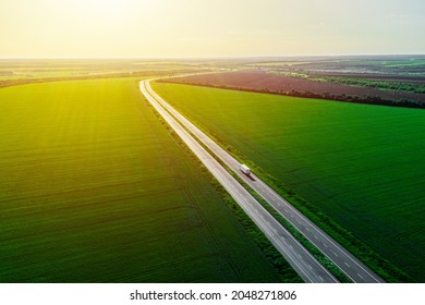 Cargo Delivery At Sunset. White Truck Driving On Asphalt Road Along The Green Fields.  Seen From The Air. Aerial View Landscape. Drone Photography. Back View.