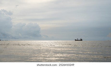 A cargo ship glides across a calm sea beneath a vast, cloud-covered sky, reflecting sunlight off the water's surface. This serene maritime scene embodies tranquility and the power of global trade. - Powered by Shutterstock