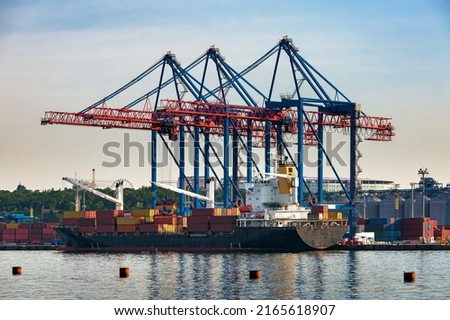 a cargo ship moored for loading at container yard with large cranes unloading goods from the deck to cargo port area. Transportation and logistics of goods by sea is industry. Sea port under sky.