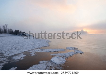 Cargo ship moving forward in front of the rising sun seen from a frozen river coast during a hazy winter dawn, Sainte-Pétronille, Island of Orleans, Quebec, Canada
