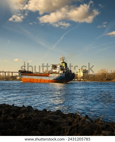 Cargo ship sailing on a river with a cityscape in the background under a sky with scattered clouds, captured from a stony riverbank.