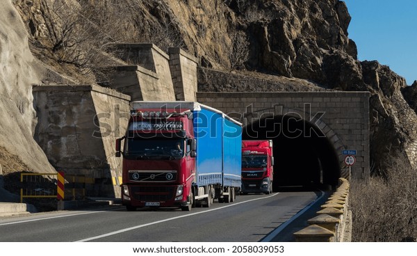Cargo trucks on the beautiful mountain road, at the\
exit of a tunnel, from below the mountain ridges. This is a Volvo\
truck. Semi-trailer carrying goods on highway. Severin, Romania,\
October 13, 2021