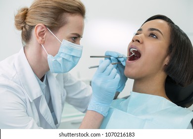 Caring about teeth. Concerned dark-haired patient sitting with her mouth opened while the dentist examining her teeth