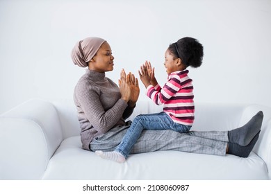 Caring african american islamic woman sitting on couch with her little daughter and clapping their hands. Pretty child playing with mother in studio with white background.