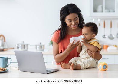 Caring African American Mother Giving Water Bottle To Her Adorable Infant Son In Kitchen, Thirsty Cute Toddler Baby Wearing Jumpsuit Sitting On Table And Enjoying Healthy Drink, Free Space