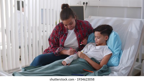 Caring African Mother Visiting Son With Broken Arm In Bed In Hospital. Happy Preteen Child Painting Arm Plaster Cast With Smiling Mom Visiting In Hospital Ward