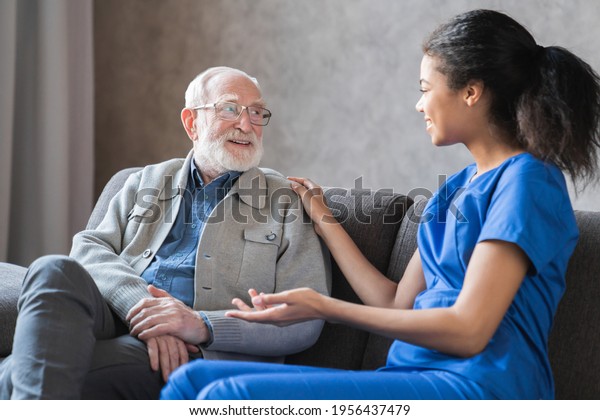 Caring african-american nurse talks to old\
elderly senior patient holds his hand sit in living room at\
homecare visit provide psychological support listen complains\
showing empathy\
encouraging.