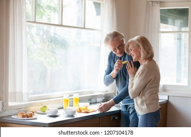 Caring Aged Husband Feed Loving Senior Wife Hugging Him From Behind, Elderly Romantic Couple Cook Breakfast At Home Together, Sensual Man Treat Beloved Woman Embracing Making Food In Kitchen