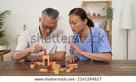 caring asian woman care attendant giving instructions while assisting elderly patient go through rehab treatment for Parkinson’s disease with building blocks at home