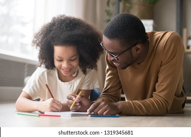 Caring black dad babysitter drawing with colored pencils teaching child girl lying on floor, african father enjoy helping kid daughter playing doing homework at home, happy family creative activity - Powered by Shutterstock