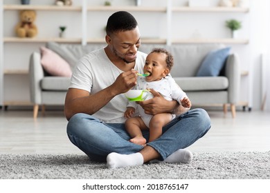 Caring Black Dad Feeding His Adorable Infant Baby From Spoon While Sitting Together On Carpet In Living Room, Young African American Father Enjoying Takic Care About Little Child, Copy Space