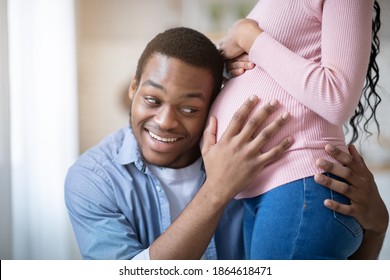 Caring Black Guy Putting His Ear On Girlfriend's Pregnant Tummy, Listening To Baby's Heartbeat At Home. Millennial African American Family Expecting Child, Expressing Love To Each Other