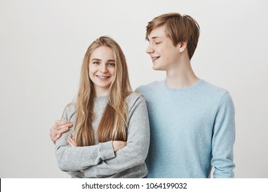 Caring boyfriend loves everything in her. Tall attractive young guy with fair hair, hugging and looking at girlfriend, smiling broadly while thinking how he love her, standing over gray wall