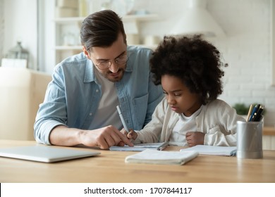Caring Caucasian father help biracial little daughter with homework at home, loving European dad and small African American girl child study together in kitchen on quarantine, homeschooling concept