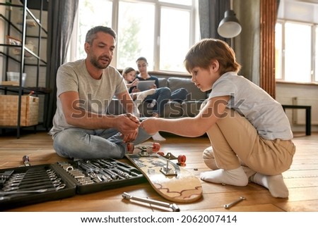 Caring Caucasian father teach teen son use deal with tools repair skateboard at home together. Young dad and small teenage boy child fix sport board with toolset equipment. Fatherhood, hobby concept.
