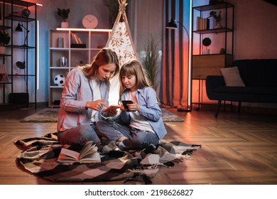 Caring Caucasian Older Sister Or Nanny With Her Younger Sister Outside Toy Wigwam Using Their Smart Phones. Dark Atmosphere At Cozy Home.