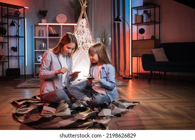 Caring Caucasian Older Sister Or Nanny With Her Younger Sister Outside Toy Wigwam Using Their Smart Phones. Dark Atmosphere At Cozy Home.