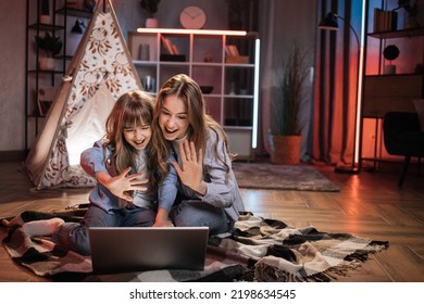 Caring Caucasian Older Sister Or Nanny With Her Younger Sister Outside Toy Wigwam Waving Their Hands While Using Laptop For Video Call. Dark Atmosphere At Cozy Home.