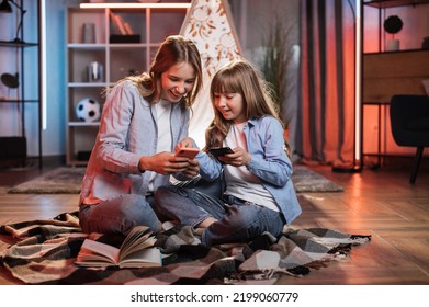 Caring Caucasian Older Sister Or Nanny With Her Younger Sister Outside Toy Wigwam Using Their Smart Phones. Dark Atmosphere At Cozy Home.