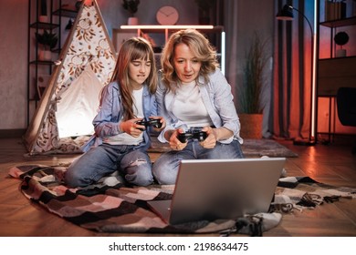 Caring Caucasian Senior Mother Or Nanny With Her Cute Daughter Girl Outside Toy Wigwam Playing Video Games Using Joysticks And Laptop. Dark Atmosphere At Cozy Home.