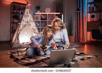 Caring Caucasian Senior Mother Or Nanny With Her Cute Daughter Girl Outside Toy Wigwam Playing Video Games Using Joysticks And Laptop. Dark Atmosphere At Cozy Home.