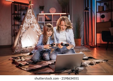 Caring Caucasian Senior Mother Or Nanny With Her Cute Daughter Girl Outside Toy Wigwam Playing Video Games Using Joysticks And Laptop. Dark Atmosphere At Cozy Home.