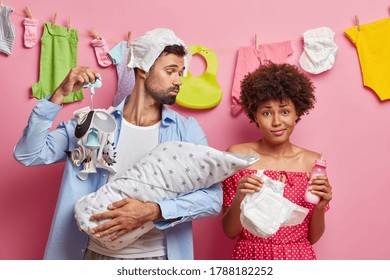 Caring Dad Looks At Baby And Shows Mobile For Falling Asleep, Enjoys Parenthood, Puzzled Curly Haired Woman Holds Diaper And Bottle Of Milk, Going To Feed Newborn And Change Nappy. Family Concept