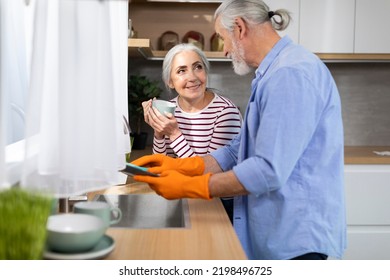 Caring Elderly Husband Helping Wife With Dishes In Kitchen, Smiling Senior Man Cleaning Plates While His Spouse Drinking Coffee, Happy Aged Spouses Sharing Domestic Chores, Free Space - Powered by Shutterstock