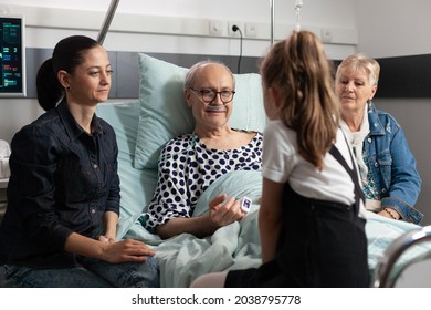 Caring Family Visiting Elderly Sick Grandfather Showing Love During Clinical Recovery In Hospital Ward. Sick Old Senior Man Resting In Bed Recovering After Medical Surgery. Heathcare Support System