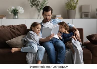 Caring father in glasses reading book to little daughter and son, hugging, sitting on cozy couch at home, family spending leisure time together, loving dad with kids engaged in educational activity