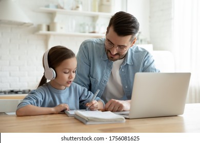 Caring Father Help Little Daughter With School Subject. Family Sit In Kitchen Table At Home Kid Girl Wears Headphones Use Laptop Makes Notes. Homeschooling Conducted By Parent, Distant E-study Concept