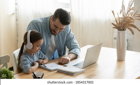 Caring father helping little daughter with school homework, sitting at table at home, child schoolgirl wearing headphones studying online, using laptop, dad checking tasks, homeschooling concept