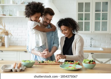 Caring father holding adorable little daughter, standing in kitchen, looking at African American mother cooking salad, shopping vegetables, multiracial family waiting for dinner or breakfast - Powered by Shutterstock