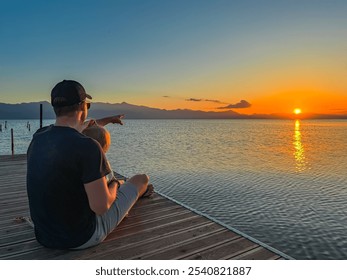 Caring father holding toddler on wooden pier on calm Skadar lake at sunset in Shkoder, Albania. Scenic view of Dinaric Alps mountains, Southern Montenegro. Perfect family vacation. Child love emotion - Powered by Shutterstock