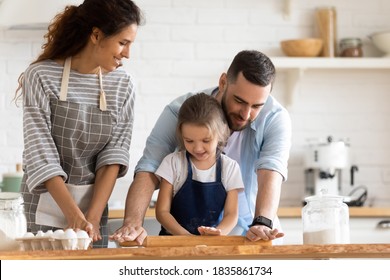 Caring father teaching little daughter wearing apron to work rolling pin, happy parents with adorable preschool girl child cooking pastry or pie, baking, enjoying leisure time together