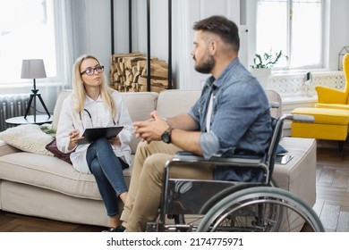 Caring Female Doctor With Medical Documents Consulting Disabled Man At Living Room. Young Male Patient In Wheelchair Listening Prescription From Therapist.