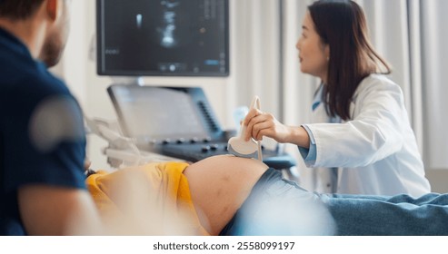 Caring Female Hospital Obstetrician Conducting an Ultrasound Exam for a Pregnant Woman. Young Couple Watching the Screen while Asian Doctor Describes the Baby’s Growth to Expectant Mother - Powered by Shutterstock