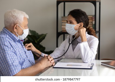 Caring Female Nurse In Medical Facemask Listen To Elderly Patient Heart With Stethoscope In Hospital. Woman GP Or Doctor In Facial Mask Do Checkup Examine Mature Man Client On Consultation In Clinic.