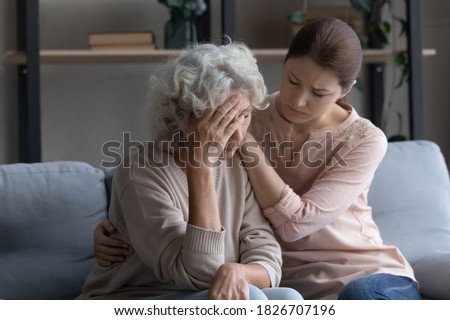 Caring grownup daughter comforting frustrated unhappy mature woman, hugging, sitting on couch at home, upset elderly female feeling unwell, touching forehead, suffering from migraine or head ache